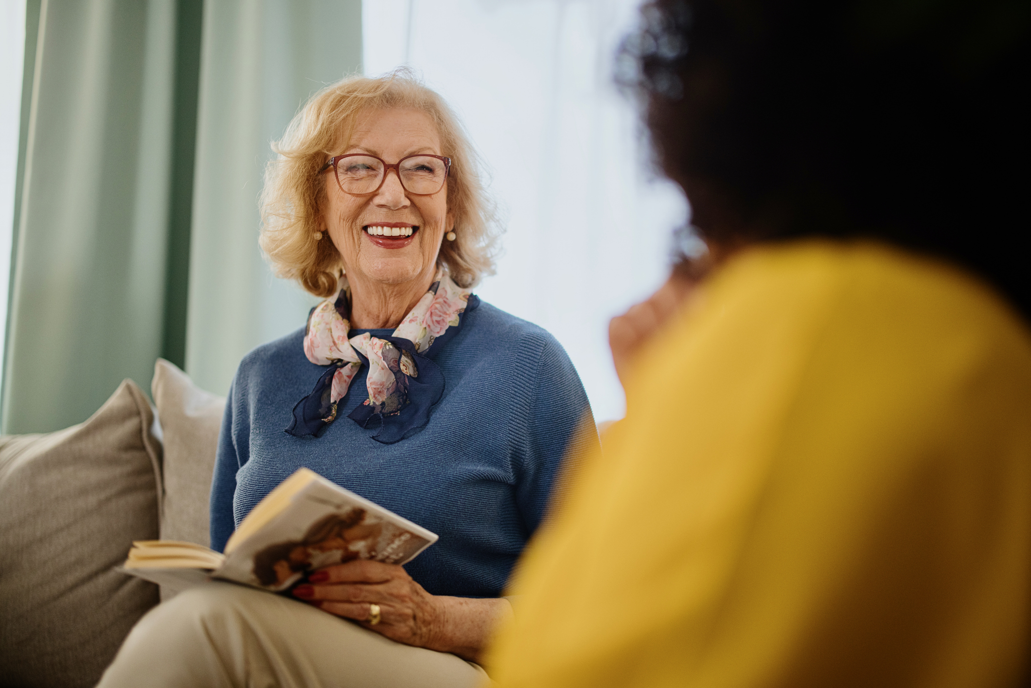 Senior woman holding book on couch smiling at another woman