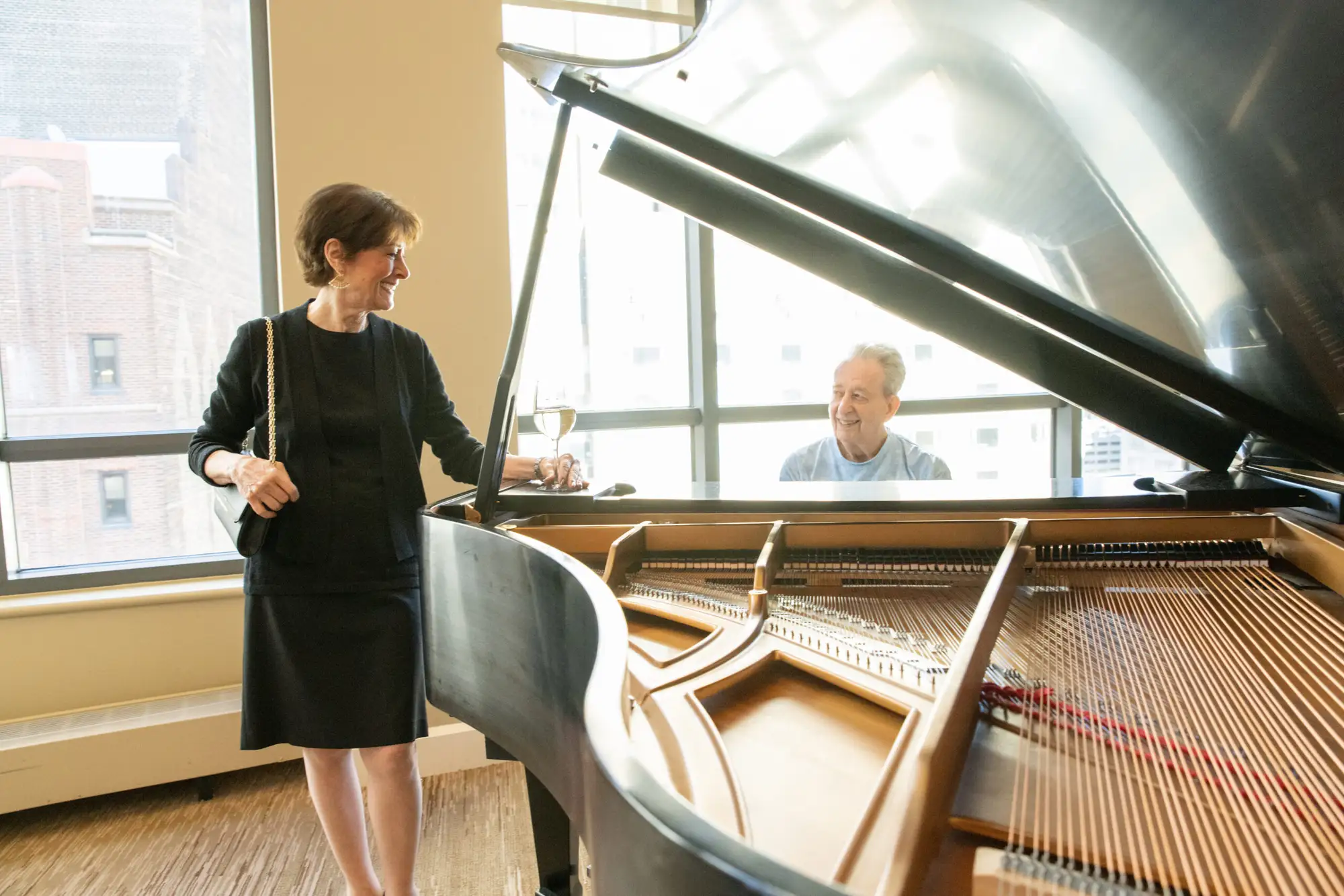 A senior man playing the piano while an elderly woman in a black dress watches, and talking about the best age to move into independent living in Chicago, IL.